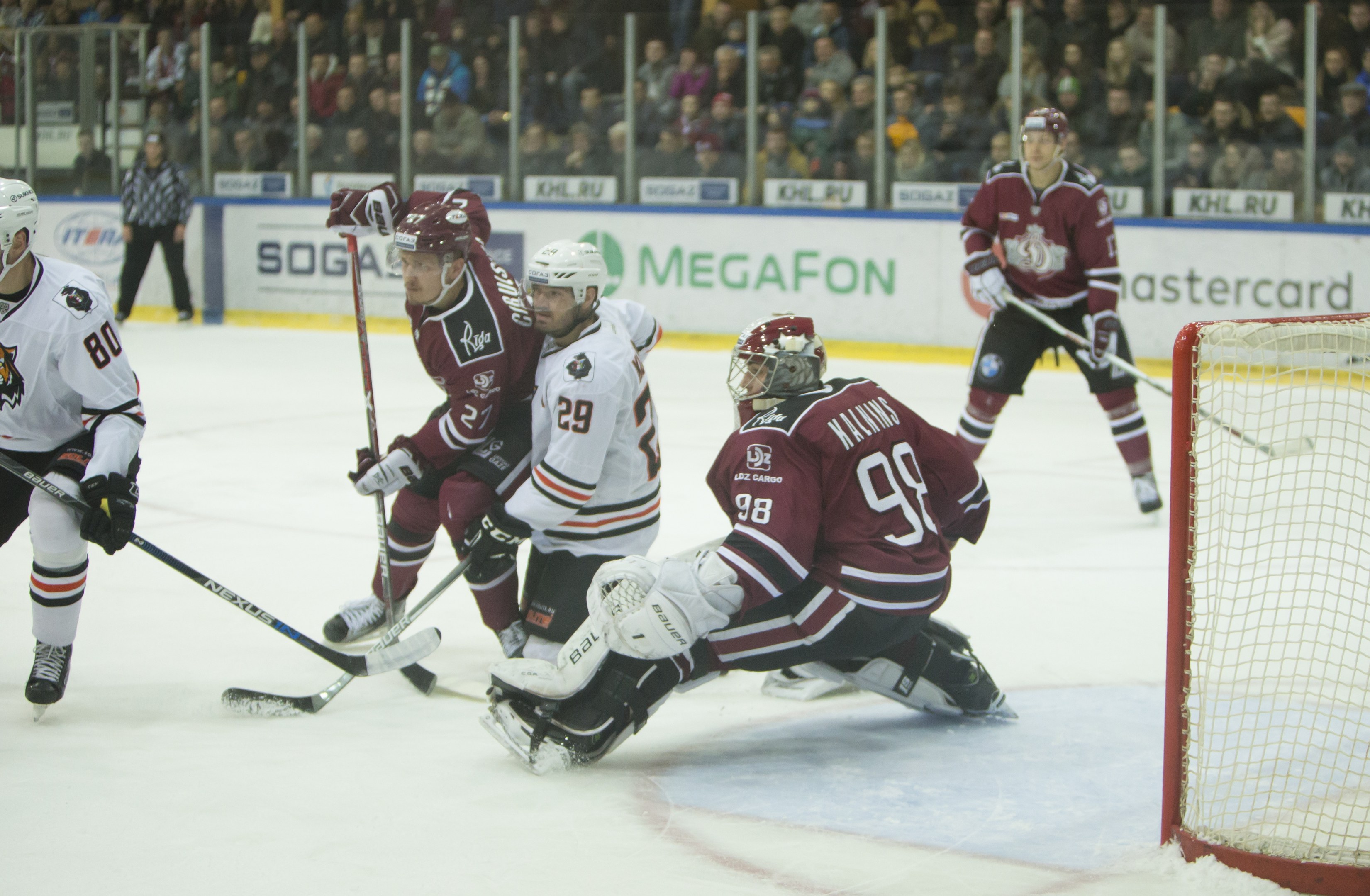 Gruppe von Menschen, die Hockey auf einem Eisstadion spielen, mit Torpfosten auf der rechten Seite, Helme tragend und Stöcke haltend, Zuschauer in den Rängen mit Bannern im Hintergrund.