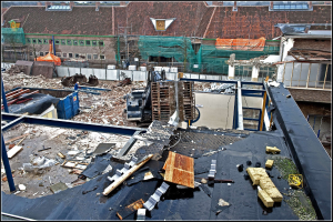 Baustelle mit einem Kran, umgeben von Gebäuden, Bäumen, Absperrungen und anderen Gegenständen, mit Text unten.