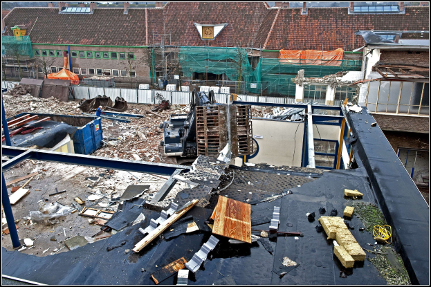 Baustelle mit einem Kran, umgeben von Gebäuden, Bäumen, Absperrungen und anderen Gegenständen, mit Text unten.