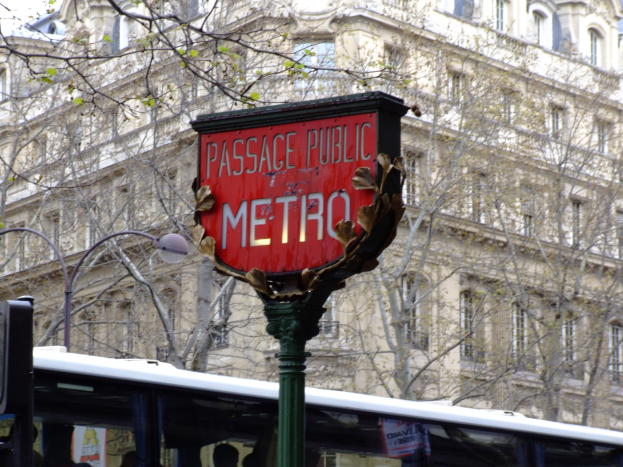 Ein Bus hält an einer öffentlichen U-Bahn-Station mit einer roten Tafel an einem Pfahl im Vordergrund, einem Gebäude mit Fenstern, Bäumen und einem klaren blauen Himmel im Hintergrund.