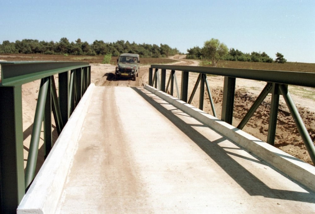 Jeep fährt über eine Brücke mit Geländern über einen Schotterweg, Bäume und einen klaren blauen Himmel im Hintergrund.