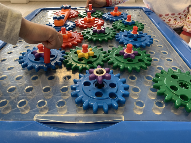 Zwei Kinder spielen mit bunten Zahnrädern auf einem Tisch, mit einer Wand im Hintergrund.