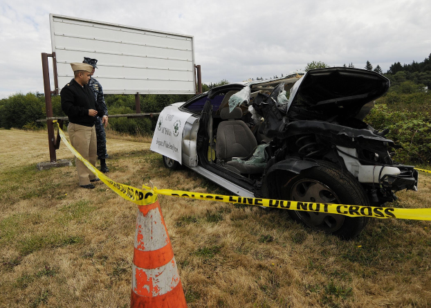 Ein Polizeibeamter in Uniform steht neben einem schwer beschädigten Auto mit eingedellter Motorhaube und zersplitterten Fenstern am Straßenrand, mit einem Verkehrskegel und Band in der Nähe, Bäumen und einem bewölkten Himmel im Hintergrund.