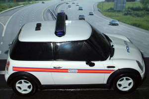 Weißer Polizeiwagen auf einer Straße außerhalb der Stadt mit Verkehr, Gras und Bäumen auf beiden Seiten.