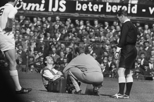 Ein Schwarz-Weiß-Foto eines Schiedsrichters, der mit einem Mann auf dem Feld spricht und von einer Gruppe von Menschen umgeben ist, mit einem Zaun, einer Fahne und Bäumen im Hintergrund.