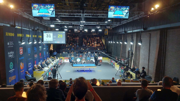 Eine große Menschenmenge sitzt vor einer Bühne mit einem Tischtennistisch in der Mitte, Stühlen auf der linken Seite, einer Wand auf der rechten Seite und Deckenlampen und Fernsehern im Hintergrund bei der Jumbo-Weltmeisterschaft 2019.