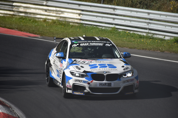 Ein BMW M4 DTM-Rennwagen auf einer Rennstrecke, umgeben von Gras, einem Zaun und Pflanzen, mit Text und Zahlen auf der Karosserie und einem sichtbaren Rad.