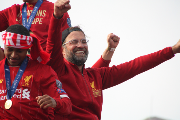 Liverpool-Trainer Jurgen Klopp feiert den Sieg der UEFA Champions League mit seinen Spielern, während er eine rote Jacke trägt und von zwei medaillen tragenden Personen umgeben ist. Der Himmel ist im Hintergrund zu sehen.