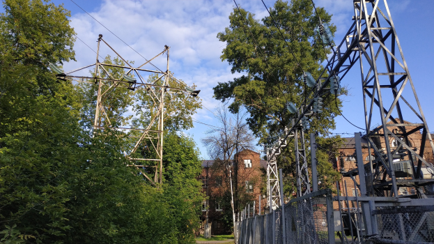 Gro├če Stromleitung durch einen Wald mit B├Ąumen, Masten, Kabeln und einem Zaun, mit im Hintergrund sichtbaren Geb├Ąuden unter einem bew├Âlktem Himmel.