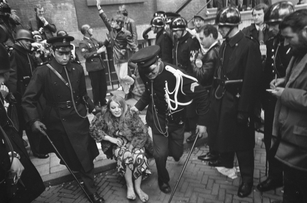 Schwarz-weißes Bild einer Frau, die von der Polizei durch eine Menge mit Helmen und Stöcken geführt wird, vor einem Gebäude mit Treppe und Geländer im Hintergrund.