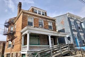 Eine Reihe von baufälligen Häusern an einer Straßenecke mit zerbrochenen Fenstern, bröckelnden Wänden, Geländern, Stufen, einem Zaun, einer Satellitenschüssel, Kabeln und einem bewölkten Himmel im Hintergrund.