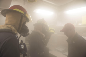 Eine Gruppe von Feuerwehrleuten in Helmen, mit Rauch aus dem Mund, einer hält eine Kamera, in einem Raum mit Lampen und anderen Gegenständen während einer Übung.