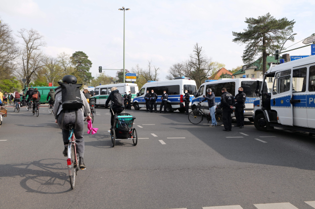 Eine Gruppe von Menschen, die auf Fahrrädern eine Straße entlangfahren, mit Polizeiwagen und anderen Fahrzeugen, einige tragen Helme und haben Taschen oder schieben Kinderwagen, mit Bäumen, Gebäuden und Verkehrsschildern im Hintergrund.