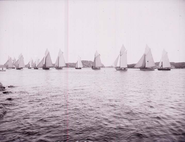 Schwarzes und weißes Foto von Segelbooten auf dem Wasser mit Hügeln im Hintergrund, klarem Himmel und 'New York Yacht Club'-Text unten.