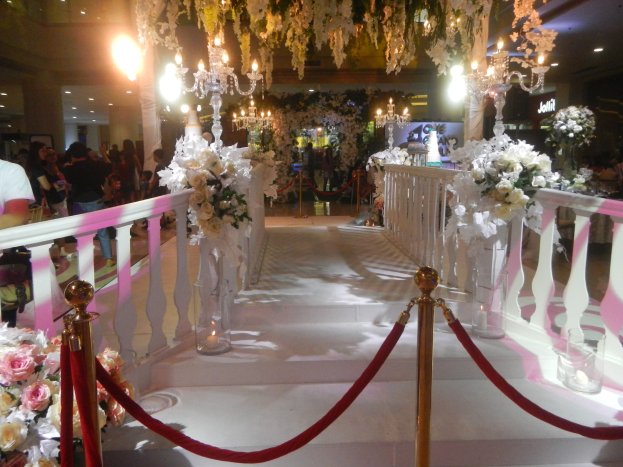 Eine Hochzeitszeremonie in einem Einkaufszentrum mit weißem Teppich, dekorativen Geländern, Blumenschmuck, Kronleuchtern und Deckenbeleuchtung, bei der zahlreiche Gäste anwesend sind, von denen einige Taschen tragen.