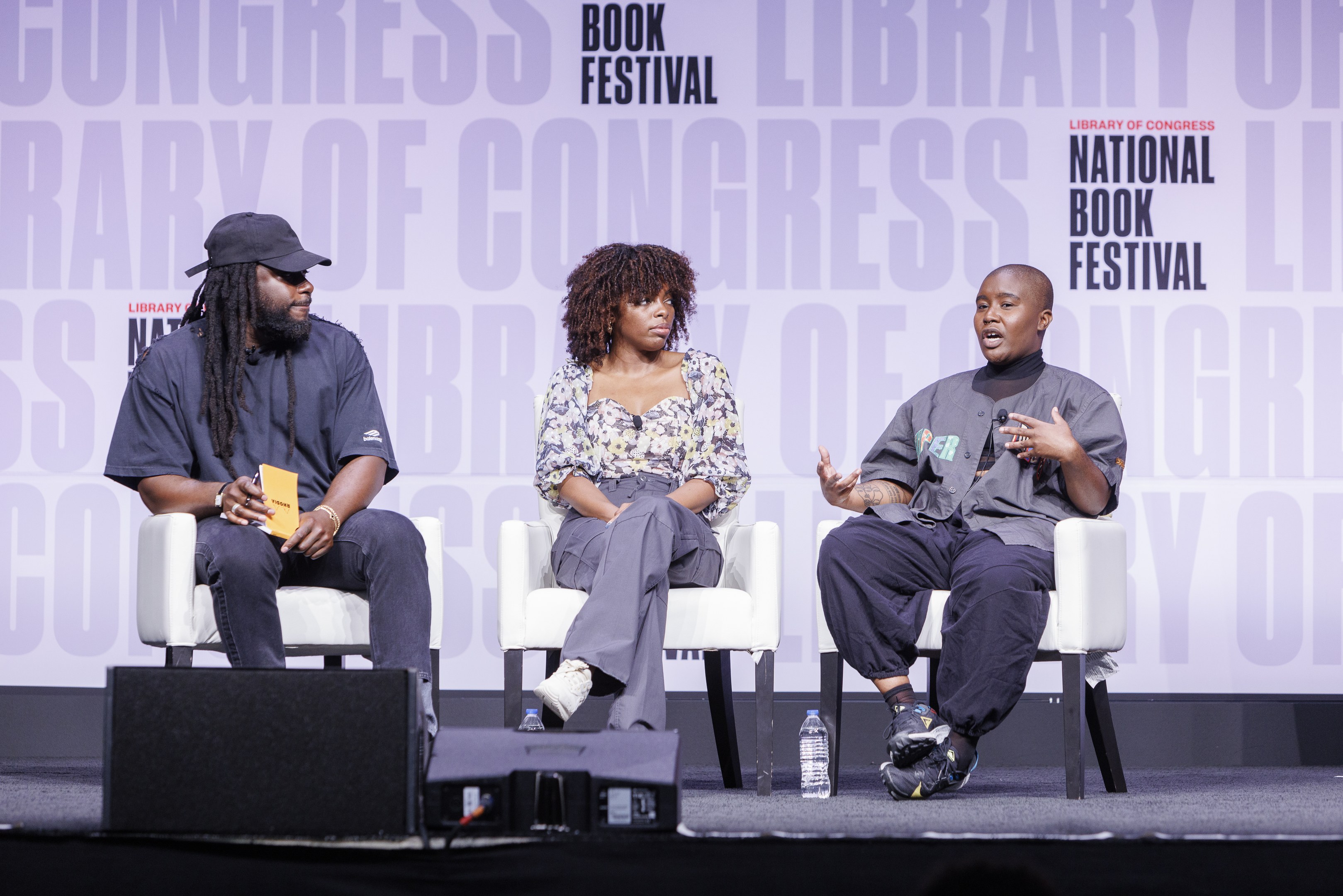 Drei Personen sitzen auf Stühlen auf einer Bühne vor einem Bildschirm, wobei ein Mann links ein Buch hält, eine Frau in der Mitte ein Mikrofon hält und verschiedene Gegenstände auf der Bühne stehen, einschließlich Flaschen, mit einem Banner im Hintergrund, auf dem 'Bibliothekskongress-Buchfestival' steht.