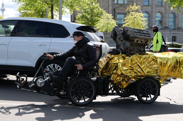 Ein Mann im Rollstuhl mit einem großen Motor auf dem Rücken, umgeben von Fahrzeugen auf einer Straße mit Bäumen, Gebäuden, Polen und einem klaren blauen Himmel im Hintergrund, trägt eine schwarze Jacke und eine Mütze und hält ein Objekt.