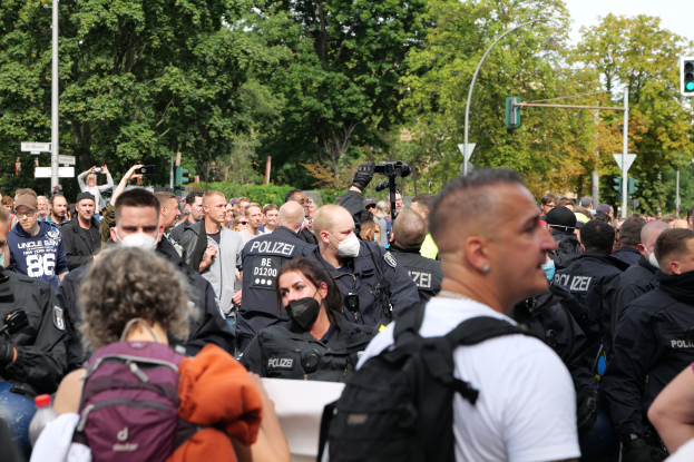 Eine große Gruppe von Menschen steht vor einer Menge von Polizeibeamten, einige tragen Masken und Rðcken, andere halten Kameras, mit Laternenmästen, Verkehrsampeln, Schildern, Bäumen und einem klaren blauen Himmel im Hintergrund.