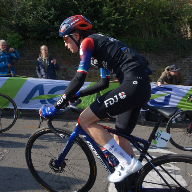 Professionelle Radrennfahrerin Katie Archibald fährt ein Fahrrad auf einer Straße, trägt einen Helm, eine Schutzbrille, Handschuhe und Schuhe und hat eine Menschenmenge und ein Banner im Hintergrund.