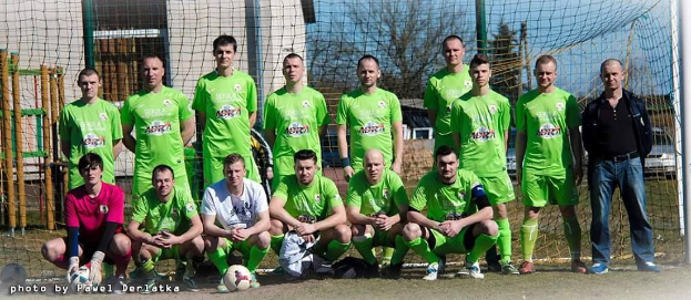 Eine Gruppe von Männern in grünen T-Shirts steht und hockt auf einem Fußballfeld mit einem Ball davor, einem Torpfosten rechts daneben und einem Hintergrund aus Bäumen, Gebäuden, Pfosten, Fahrzeugen und dem Himmel.