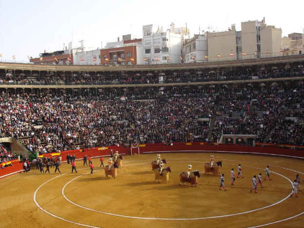 Menschen gehen auf dem Boden, einige reiten Pferde, andere sitzen im Stadion und schauen zu, Flaggen und Gebäude sind im Hintergrund unter dem Himmel zu sehen.