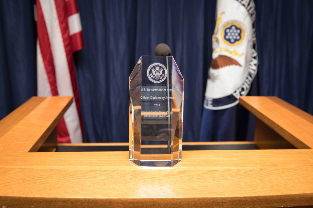 Ein Glaspreis auf einem Holzpodest mit einem Mikrofon davor, Flaggen und Vorhänge im Hintergrund.