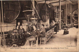 Ein Schwarz-Weiß-Foto von Frauen in einer Fabrik, mit einigen sitzend und anderen stehend zwischen Maschinen, und Text unten lesend "Amer Picon - Use de Bordeaux-Bastide".