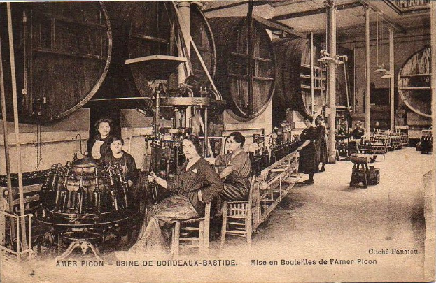 Ein Schwarz-Weiß-Foto von Frauen in einer Fabrik, mit einigen sitzend und anderen stehend zwischen Maschinen, und Text unten lesend "Amer Picon - Use de Bordeaux-Bastide".