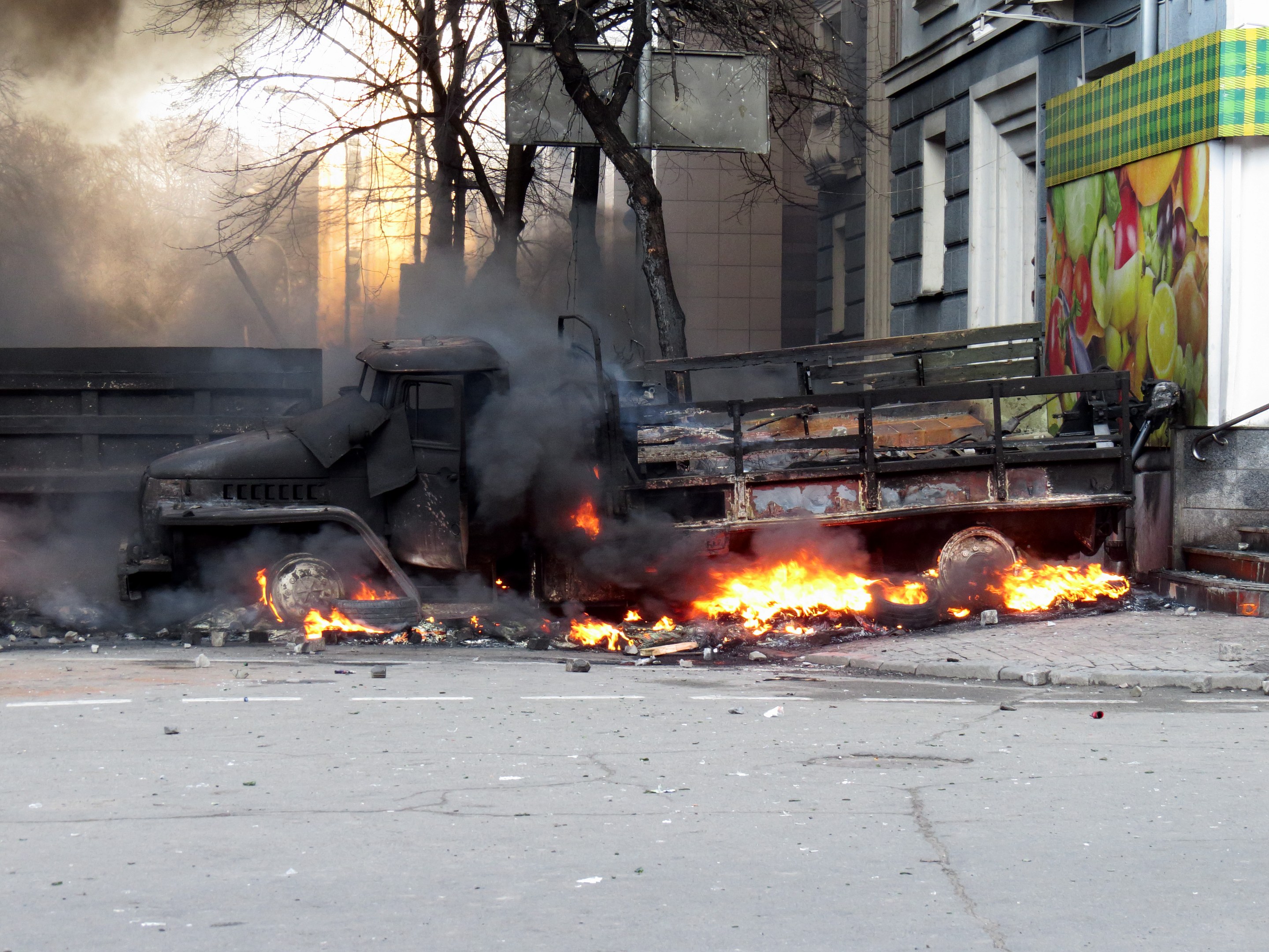 Ein großer Lkw brennt am Straßenrand mit dicken schwarzen Rauchwolken, Bäume und Gebäude sind im Hintergrund unter einem klaren Himmel zu sehen.