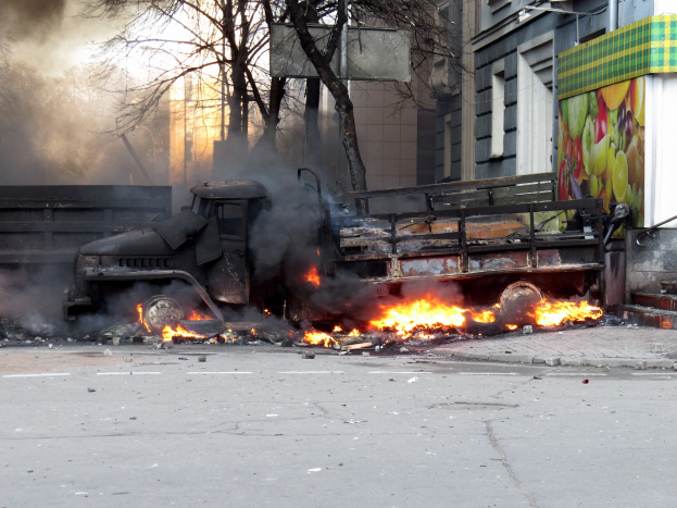 Ein großer Lkw brennt am Straßenrand mit dicken schwarzen Rauchwolken, Bäume und Gebäude sind im Hintergrund unter einem klaren Himmel zu sehen.