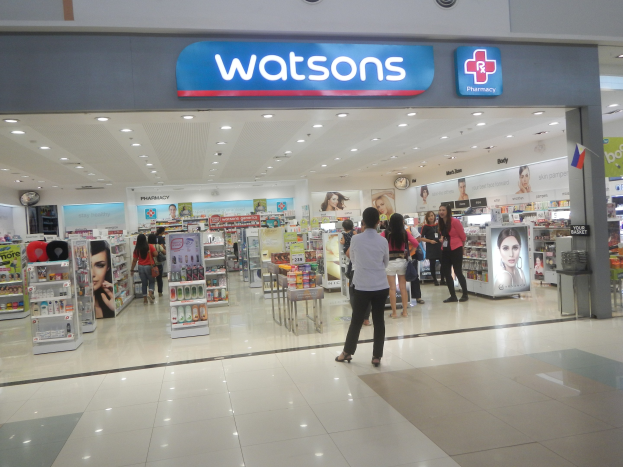 Außenansicht einer Watsons Apotheke in einem Einkaufszentrum mit Menschen, Regalen, Schildern, einer Wanduhr und Deckenleuchten.