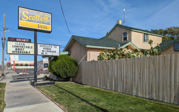 Ein schottisches Motel in San Francisco, Kalifornien, mit einem Haus, einem Holzzaun, Pflanzen, Bäumen, Schildern, Strommasten, einer Straßenlaterne, Fahrzeugen auf der Straße und einem bewölkten Himmel.