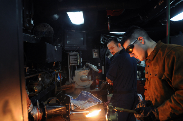 Zwei Männer mit Brille arbeiten an einem Metallstück in einer Werkstatt, einer verwendet ein Schweißgerät, während Werkzeuge und Equipment auf dem Tisch liegen; ein Fernseher und Lampen sind im Hintergrund zu sehen.