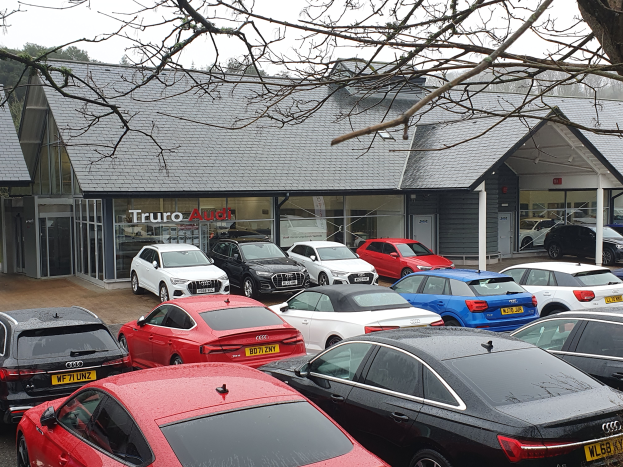 Ein Autohaus mit mehreren parkenden Autos vorne, umgeben von Bäumen unter einem klaren blauen Himmel, mit einem Gebäude mit Dach, Glaswänden und einem Schild mit Text.