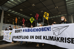 Eine Gruppe maskierter Personen mit einem Banner "Sturzflug in die Klimakrise" steht vor einem gläsernen Gebäude mit Säulen, beleuchtet von Deckenlampen.