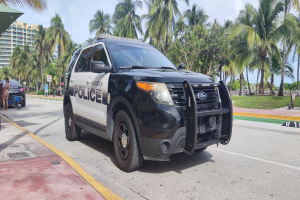 Polizeiauto am Straßenrand mit Menschen, Fahrrädern, Gras, Bäumen, Gebäuden und einem klaren blauen Himmel im Hintergrund; Vorderrad sichtbar.