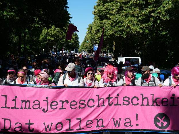 Eine Gruppe von Menschen marschiert mit einem pinken Banner mit der Aufschrift 'Frauenmarsch in Berlin' die Straße entlang, einige tragen Mötzen und Schutzbrillen, andere halten Fahnen, vor dem Hintergrund von Bäumen, Fahrzeugen und Himmel.