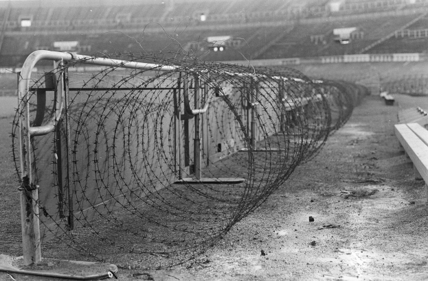 Schwarzes und weißes Foto eines Stacheldrahtzauns vor einem Stadion, mit Bänken auf der rechten Seite und einem Stadion im Hintergrund.