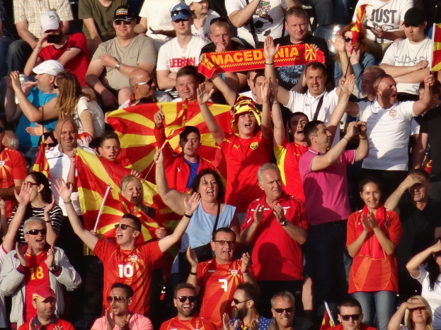 Eine Menge im Stadion hält ihre Hände hoch, einige tragen Mützen und Schutzbrillen, halten Fahnen und Schilder mit Text, mit Geländern im Hintergrund.