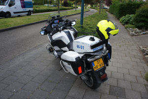 Eine Polizei-Motorrad auf dem Seitenstreifen geparkt mit einem Helm darauf, mit Bäumen, Pfählen, Gebäuden, Pflanzen, Gras, Steinen und anderen Fahrzeugen im Hintergrund.