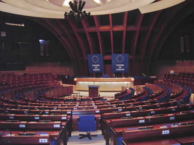 Großer halbrunder Konferenzraum im Europäischen Parlament mit Tischen, Stühlen, einem zentralen Podium und Texttafeln an den Wänden unter einer beleuchteten Decke.