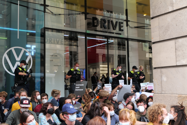 Eine große Gruppe maskierter Menschen mit Schildern vor einem Glasgebäude mit Text, einige tragen Mützen und halten Kameras, was auf eine Demonstration in Melbourne, Australien, hinweist.