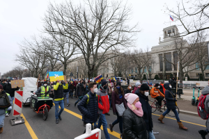 Eine große Gruppe von Menschen nimmt an einer Protestdemo auf einer Straße in Washington, D.C. teil, wobei einige Schilder und Banner halten, andere Fahrräder fahren und Schilder, Bäume und ein klarer blauer Himmel im Hintergrund zu sehen sind.