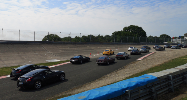 Eine Gruppe von Autos, die auf einer Rennstrecke fahren, umgeben von Verkehrskegeln, Gras, einem Zaun, Bäumen, Gebäuden, Flaggen und einem bewölkten Himmel.