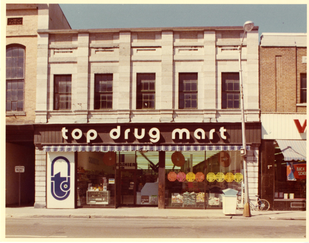 Eckladen namens "Top Drug Mart" mit einem Schild, Fenstern, einem Straßenschild, einem geparkten Fahrrad, einem Müllcontainer und einem bewölkten Himmel.