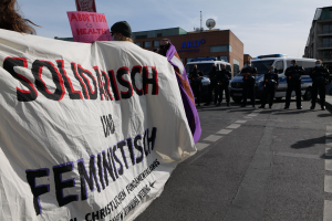 Eine Gruppe von Menschen marschiert auf einer Straße, hält ein Banner mit der Aufschrift "Solidarität und Feminismus" und hat parkende Fahrzeuge und Gebäude im Hintergrund unter einem klaren blauen Himmel.