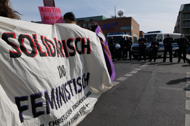 Eine Gruppe von Menschen marschiert auf einer Straße, hält ein Banner mit der Aufschrift "Solidarität und Feminismus" und hat parkende Fahrzeuge und Gebäude im Hintergrund unter einem klaren blauen Himmel.