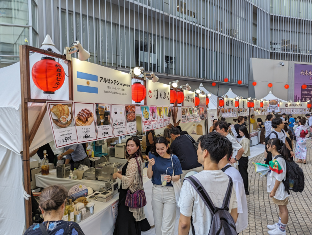 Eine Gruppe von Menschen auf einem Essensfest in Japan versammelt sich um einen Tisch mit Essen, einige halten Gläser und tragen Taschen, während andere die Gegenstände auf dem Tisch untersuchen.