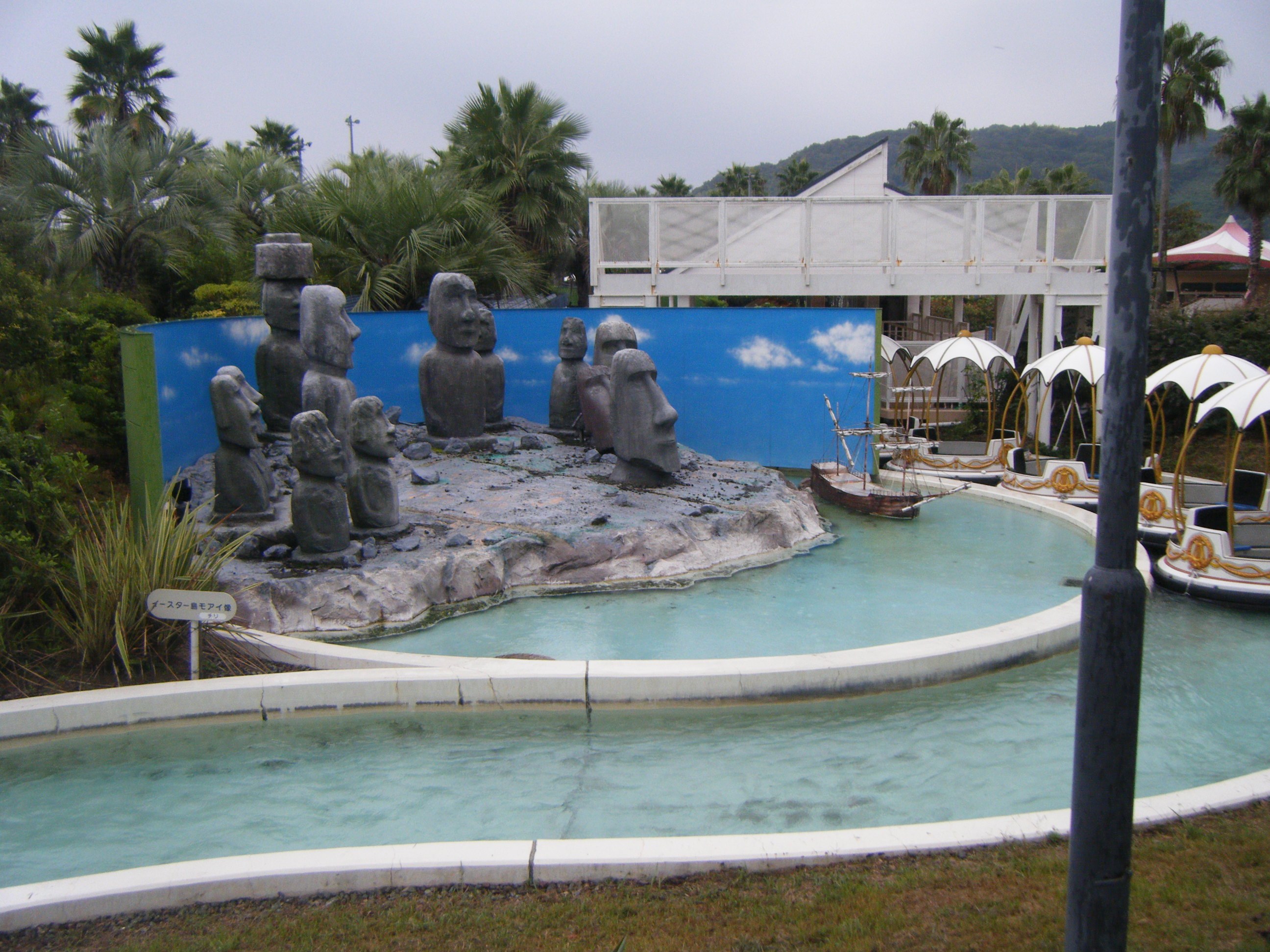 Miniatur-Golfplatz mit einem zentralen Brunnen umgeben von Statuen, Wasser, Rasen, Pflanzen, Bäumen, einem Pfahl, Zelten, einer Brücke, einem Gebäude und fernen Hügeln unter einem klaren blauen Himmel.
