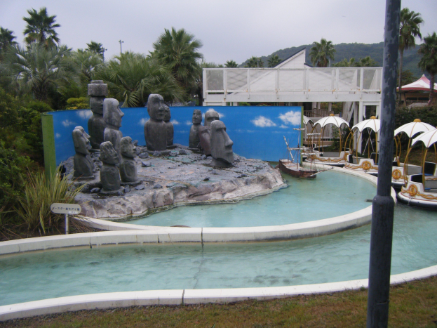 Miniatur-Golfplatz mit einem zentralen Brunnen umgeben von Statuen, Wasser, Rasen, Pflanzen, Bäumen, einem Pfahl, Zelten, einer Brücke, einem Gebäude und fernen Hügeln unter einem klaren blauen Himmel.